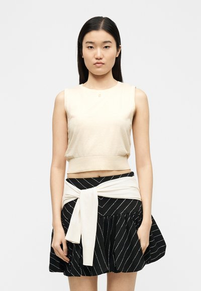 Γυναίκα με μακριά σκούρα μαλλιά φοράει αμάνικο μπεζ τοπ και μαύρη φούστα με λευκές διαγώνιες ρίγες, στέκεται μπροστά σε απλό λευκό φόντο.