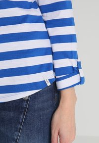 Blau-weiß gestreiftes Langarmhemd mit umgeschlagenen Ärmeln und einem kleinen Etikett. Getragen mit dunklen Denim-Jeans. Aus weichem Baumwollstoff.