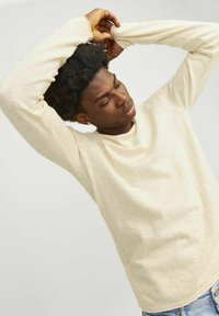 Camisa de manga larga beige claro con un acabado texturizado, cuello redondo y un corte ligeramente holgado, combinada con unos pantalones vaqueros de denim claros.