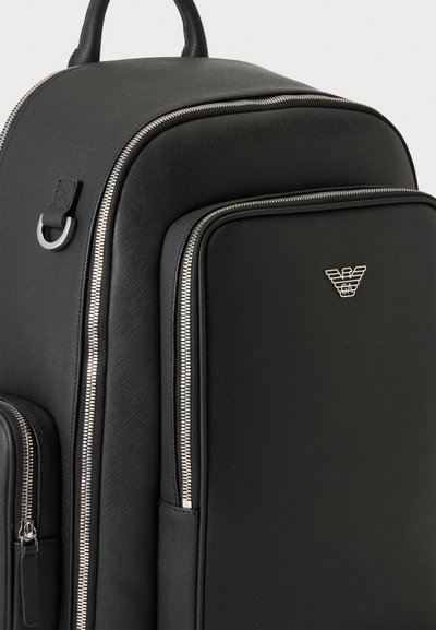 Sac à dos en cuir noir avec des fermetures éclair argentées et un logo aigle Armani en métal sur la poche avant, présenté sur un fond uni clair.
