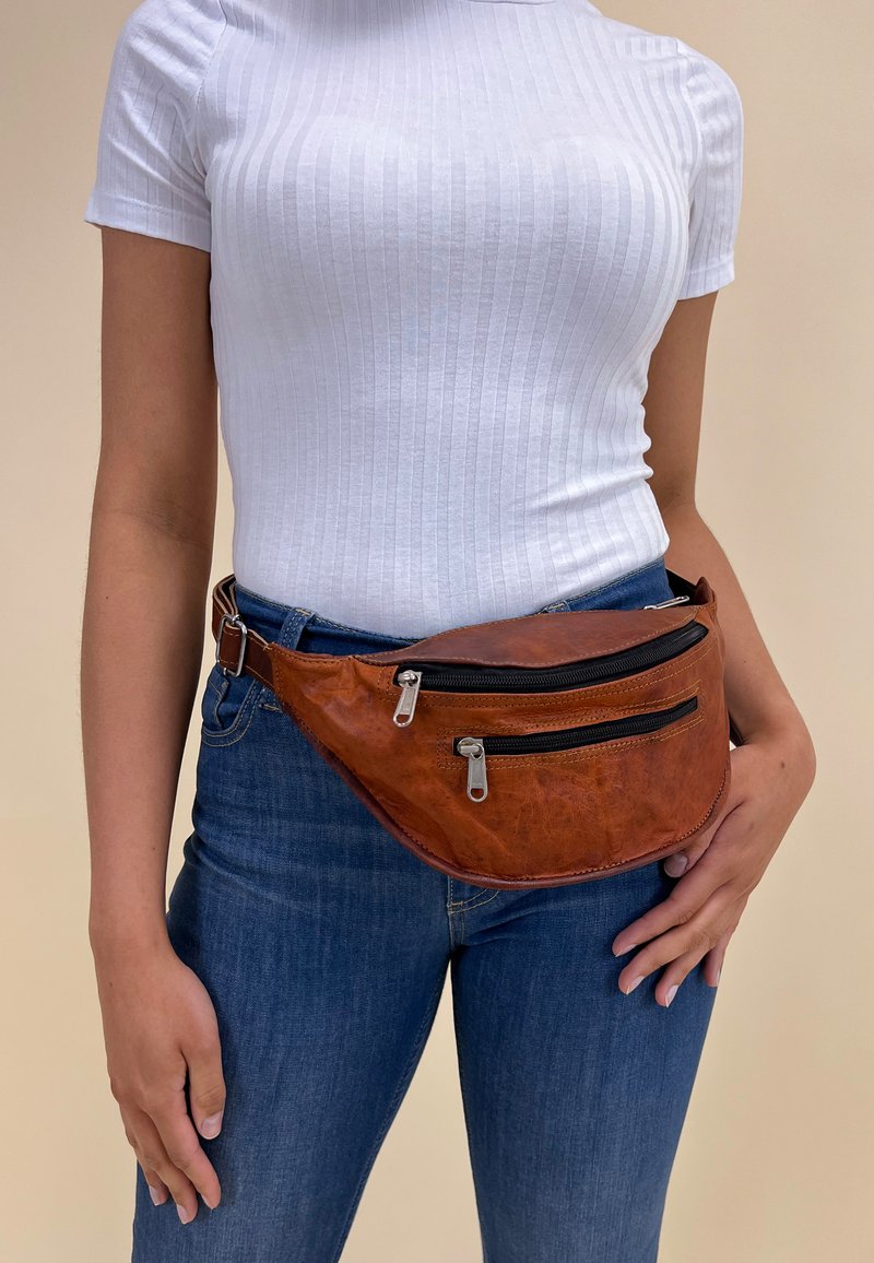 Ruda odinė fanny pack'as su dviem juodais užtrauktukais, išlenkta forma ir reguliuojamu diržu, dėvimas ant baltos griovelės viršaus ir mėlynų džinsų.