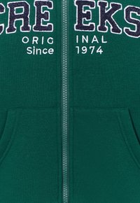 Sudadera con cremallera verde con texto bordado en blanco y marino. Material suave y texturizado, con una cremallera central y bolsillos frontales.