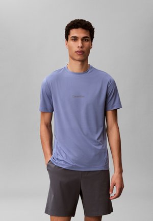 Jonge man met krullend donker haar, gekleed in een lichtblauw Calvin Klein T-shirt en donkergrijze korte broek, staande tegen een effen achtergrond.