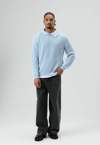 Man met gevlochten haar, gekleed in een lichtblauwe gebreide trui, zwarte wijde broek en zwarte schoenen, staand tegen een effen grijze achtergrond.