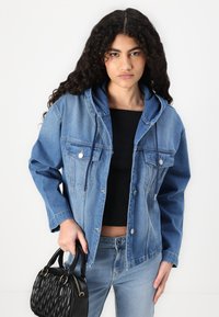 Τζάκετ από τζιν σε ανοιχτό μπλε χρώμα με κουκούλα, μπροστινές τσέπες και κουμπιά με κλιπ. Φοριέται πάνω από ένα μαύρο μπλούζα, συνδυασμένο με μπλε τζιν και μια μαύρη τσάντα.