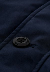 Marineblauer Stoff im Nahaufnahme mit einem großen schwarzen Knopf mit vier Löchern, geprägtem Text "TOM TAILOR REG." und dem Jahr "1962". Glatte Textur.