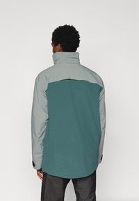 Férfi, aki magas galléros, hosszú ujjú, szürke és türkiz színű, két tónusú kabátot visel, háttal állva egy egyszínű világos háttér előtt.