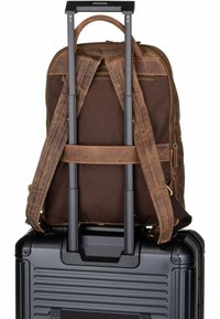 Sac à dos en cuir marron attaché à une valise noire à roulettes avec des poignées métalliques, présentant des détails de couture et des bretelles ajustables.