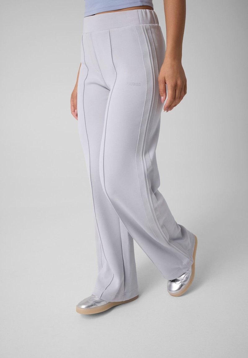 Personne portant un pantalon large gris clair avec des coutures verticales et des chaussures argentées slip-on sur un fond gris uni.