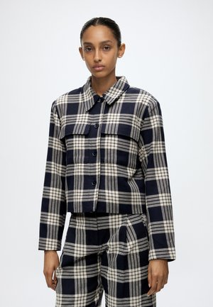 Donna che indossa una giacca a quadri blu navy e bianca con bottoni e pantaloni in tinta coordinata, in piedi davanti a uno sfondo chiaro uniforme.