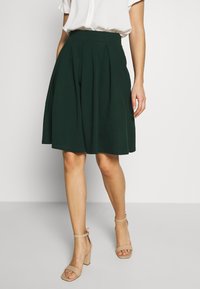 Donna in una gonna plissettata verde scuro al ginocchio, camicetta bianca a maniche corte e sandali con tacco a blocco beige con cinturino alla caviglia.