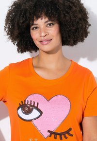 Orange T-shirt med et pink hjerte design med palietter, en broderet øje, og sorte øjenvipper detaljer. Korte ærmer og rund halsudskæring.
