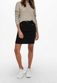 Jupe en jean noire avec poches à zip avant, coupe ajustée, longueur mi-cuisse, associée à un pull beige en maille et des baskets blanches.