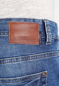 I jeans in denim blu presentano una toppa in pelle marrone con la scritta "Camel Active" in rilievo, con dettagli di cucitura lungo la vita e le tasche.