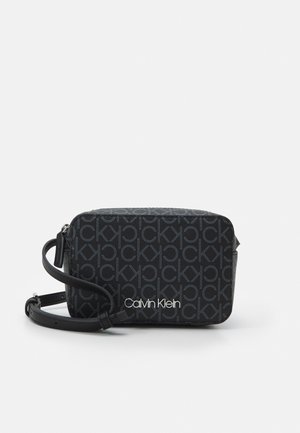 Černá designová kabelka přes rameno s texturovaným vzorem loga, vybavená zipovým uzávěrem a nastavitelným popruhem. Bílé označení značky "Calvin Klein".