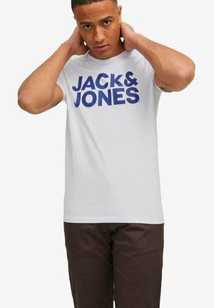 Jeune homme ajustant son col, portant un t-shirt blanc Jack & Jones et un pantalon foncé sur un fond uni.