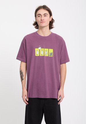 Jeune homme aux cheveux mi-longs portant un t-shirt violet avec quatre panneaux jaunes montrant une séquence de cassure d’un vase, associé à un pantalon noir.