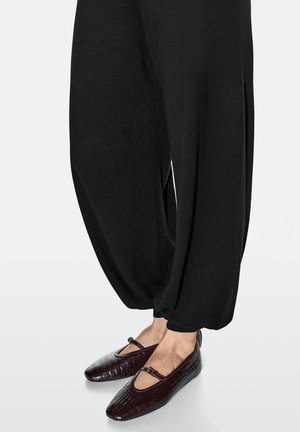 Schwarze, locker sitzende Hose aus weichem Stoff mit zulaufenden Knöcheln, kombiniert mit burgunderfarbenen, krokodilprägemusterten Ballerinas mit einem Riemen über dem Fuß.