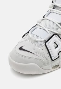 Witte sportieve schoen van mesh en leer, met een prominent zwart "A" accent, een gestructureerd paneel en een zilveren Nike swoosh-logo.