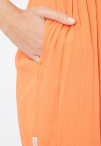 Tela naranja con una apariencia suave y texturizada, que presenta un bolsillo lateral y una pequeña etiqueta de marca visible en el dobladillo.