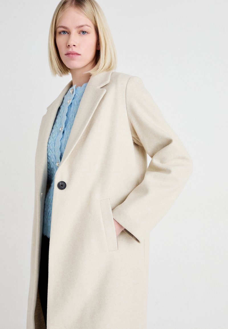 Beige Wollmantel mit Reverskragen, Ein-Knopf-Verschluss und Seitentaschen, über einem blauen Kabelstrickpullover getragen. Glatte Textur.