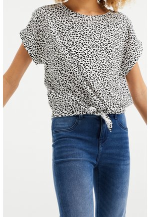Femme portant un haut blanc à manches courtes imprimé léopard noué à la taille et un jean skinny bleu taille haute sur fond blanc.
