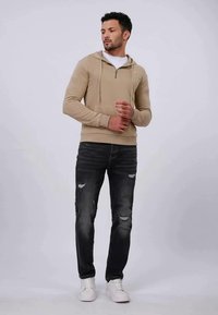 Felpa zip-up beige con tasca a canguro, abbinata a jeans neri strappati e sneakers bianche. Tessuto liscio e design aderente.