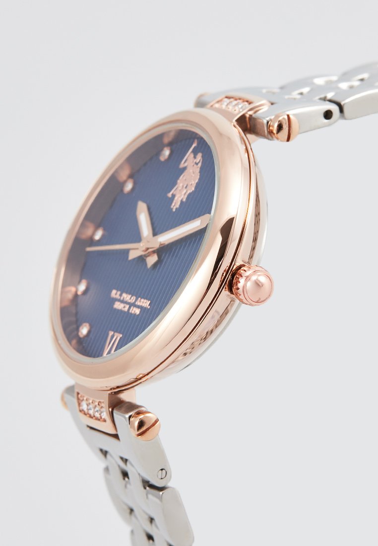 Reloj rosa dorado con una esfera texturada azul marino, pulsera de eslabones plateados y logotipo distintivo. Las características incluyen marcadores horaria y una corona.