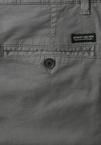 Poche arrière d'un pantalon gris pour homme avec fermeture à bouton et étiquette en tissu "Street One Men" cousue au-dessus de la poche.