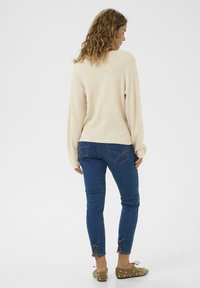 Beige Pullover mit langen Ärmeln, kombiniert mit blauen Skinny-Jeans, die Schleifenakzente an den Bündchen aufweisen. Braune Schuhe mit weißen Punkten.