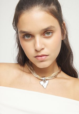 Joven con cabello castaño lleva blusa blanca con hombros descubiertos y collar beige con un gran colgante de corazón plateado, fondo neutro.