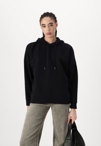 Μαύρη κουκούλα φόρμα με χαλαρή εφαρμογή, κορδόνια και μακριά μανίκια, σε συνδυασμό με ανοιχτό γκρι παντελόνι και κρατώντας μια μαύρη τσάντα.