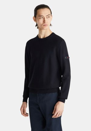 Młody mężczyzna z ciemnymi, związanymi do tyłu włosami nosi czarną bluzę z długim rękawem i granatowe spodnie, patrzy w bok na jednolitym białym tle.