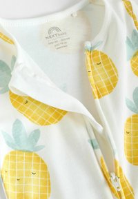Vit zip-up baby onesie med gula ananastryck och rutmönster, gröna blad och en mjuk textur. Etikett synlig inuti.