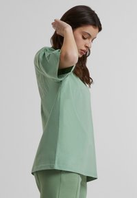 Žalia trumpa rankovė t-shirt, pagaminta iš minkštos medvilnės, su laisvu siluetu; turi šoninius įpjovimus ir subtilius reljefinius ornamentus ant audinio.