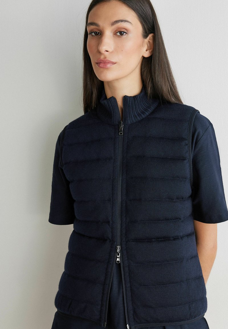 Kobieta o długich ciemnych włosach, ubrana w granatową pikowaną kamizelkę bez rękawów na ciemnej koszulce z krótkim rękawem, na neutralnym jasnym tle.