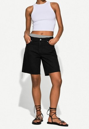 Person iført en ærmeløs hvid crop top, sorte høj-taljede knælange shorts med stribet linning og sorte flade snøresandaler.