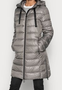 Kobieta ubrana w długi, srebrnoszary pikowany płaszcz puchowy z zamkiem z przodu, kapturem, czarnymi sznurkami ściągającymi i bocznymi detalami na zamek.