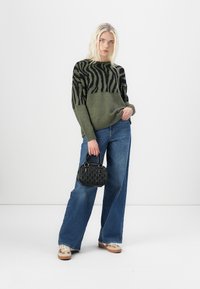 Grüner Pullover mit schwarzem Zebra-Muster, kombiniert mit weiten blauen Jeans. Hält eine kleine schwarze, strukturierte Handtasche und trägt helle Sneakers.