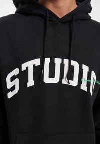 Svart hettegenser med hvit "STUDIO" tekst på brystet og grønn akse. Har snøring i hetten og en frontlomme. Myk stofftekstur.