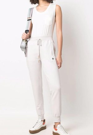 Mujer con camiseta blanca sin mangas, pantalones jogger beige claro con parche de estrella, zapatillas blancas y lleva un bolso bandolera plateado con una correa ancha.