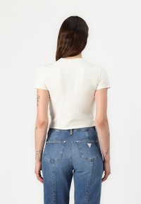 Valkoinen lyhythihainen crop-top, yhdistetty korkeavyötäröisiin sinisiin farkkuihin, joissa on kolmion muotoinen logo takataskussa.