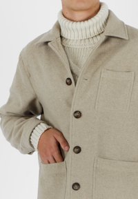 Cappotto in misto lana beige con colletto, chiusure a bottone e tasca sul petto; indossato su un maglione a collo alto crema con texture a coste.