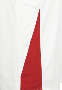 Maglietta sportiva bianca con un prominente pannello diagonale rosso sul lato, realizzata in tessuto traspirante con una superficie testurizzata.
