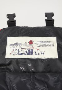 Sac à dos en tissu noir avec des accents rouges, présentant une étiquette blanche brodée représentant un design graphique et un texte sur l'histoire de la marque.