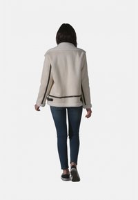 Veste en shearling couleur crème avec une texture douce, agrémentée d'accents noirs et d'un col. Portée avec un jean bleu et des baskets noires.
