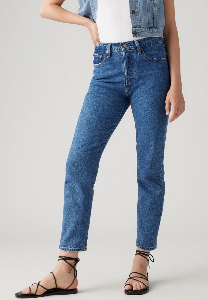 Femme portant un jean bleu taille haute, un top blanc, un gilet en jean et des sandales plates noires à brides, debout avec une main sur la hanche.