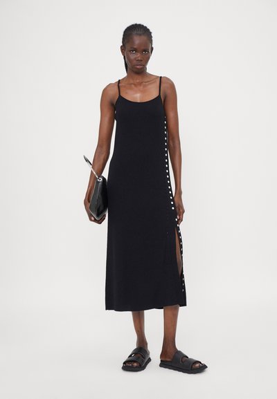 Schwarzes ärmelloses Maxikleid mit seitlichen Knopfdetails, aus weichem Stoff und gerader Silhouette, kombiniert mit einer schwarzen Clutch und Sandalen.