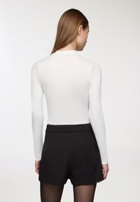 Biały długi rękaw top o dopasowanym kroju, z wysokim kołnierzem, gładką teksturą, zestawiony z czarnymi szortami o wysokim stanie i przezroczystymi czarnymi rajstopami.