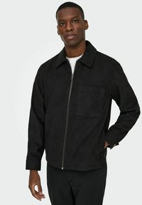 Črna velur jakna z zadrgo spredaj, koničasto ovratnikom in žepom na prsih. Dolgi rokavi s manšetami in sproščen kroj.